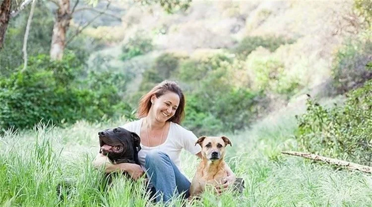 Krista Goodsitt smiling in tall green grass in the hills with two dogs, a black dog and a tan dog