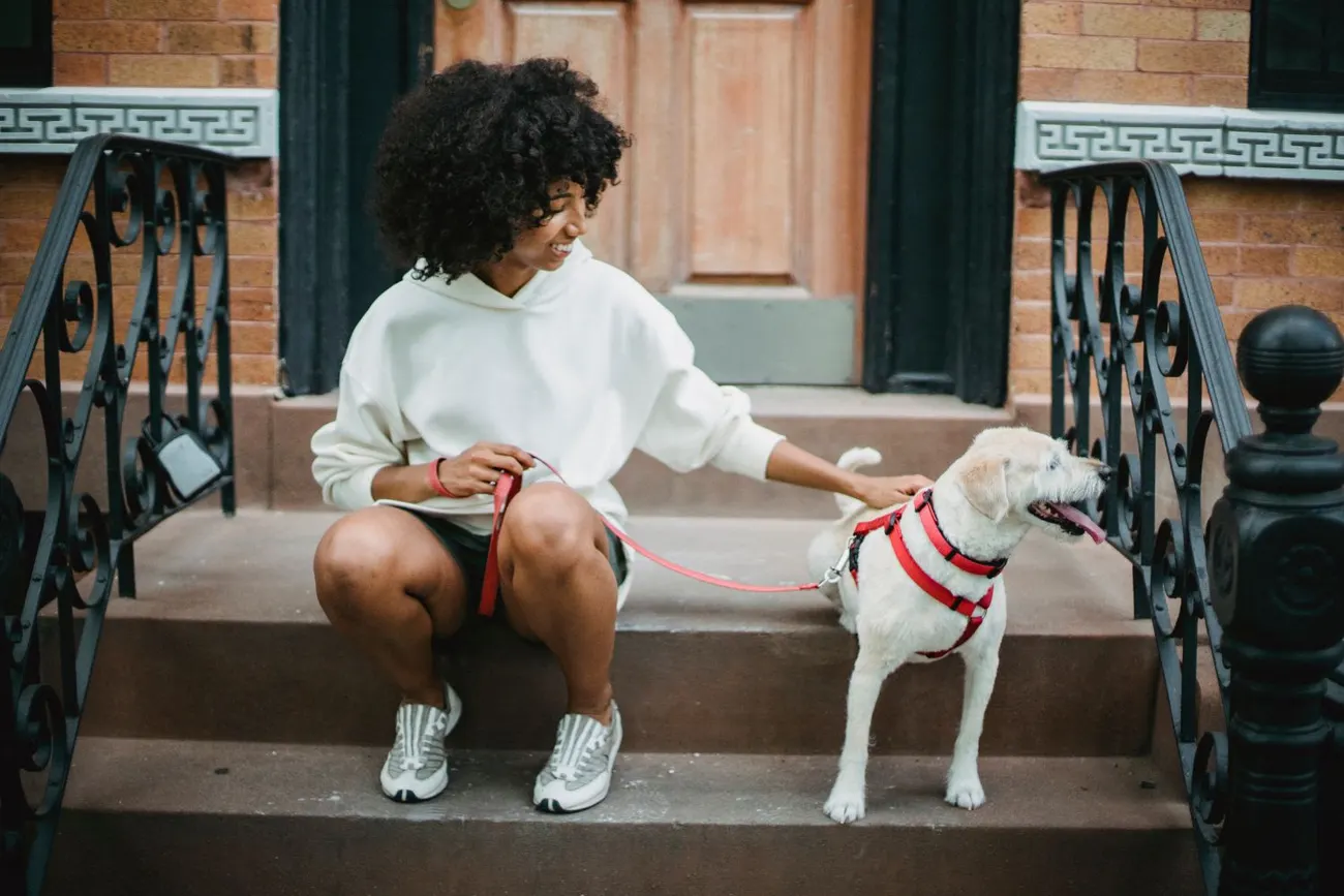 A young woman sitting on her front steps with her small white dog in a red harness